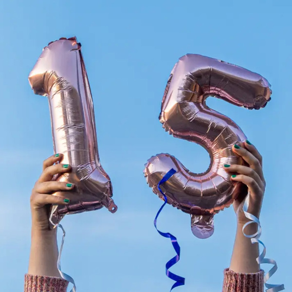 Twee handen houden roségouden folieballonnen in de vorm van het getal 15 omhoog tegen een blauwe lucht