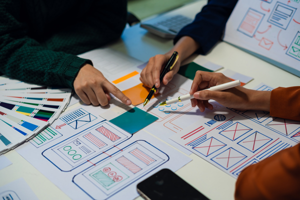 Handen met pennen wijzen naar papieren wireframes en kleurstalen van een app-ontwerp op tafel