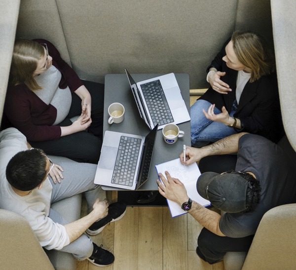Bovenaanzicht van vier collega's in overleg rond een tafeltje met laptops, koffiekopjes en een notitieblok